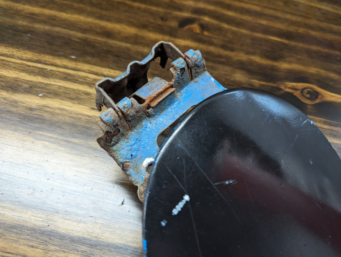 Close-up of a rusty hinge on an OEM Fuel Filler Neck Door Gas Tank Cover in Black for 2001-2004 Chevy S10 & GMC Sonoma by General Motors, resting on a wood surface with visible grain.