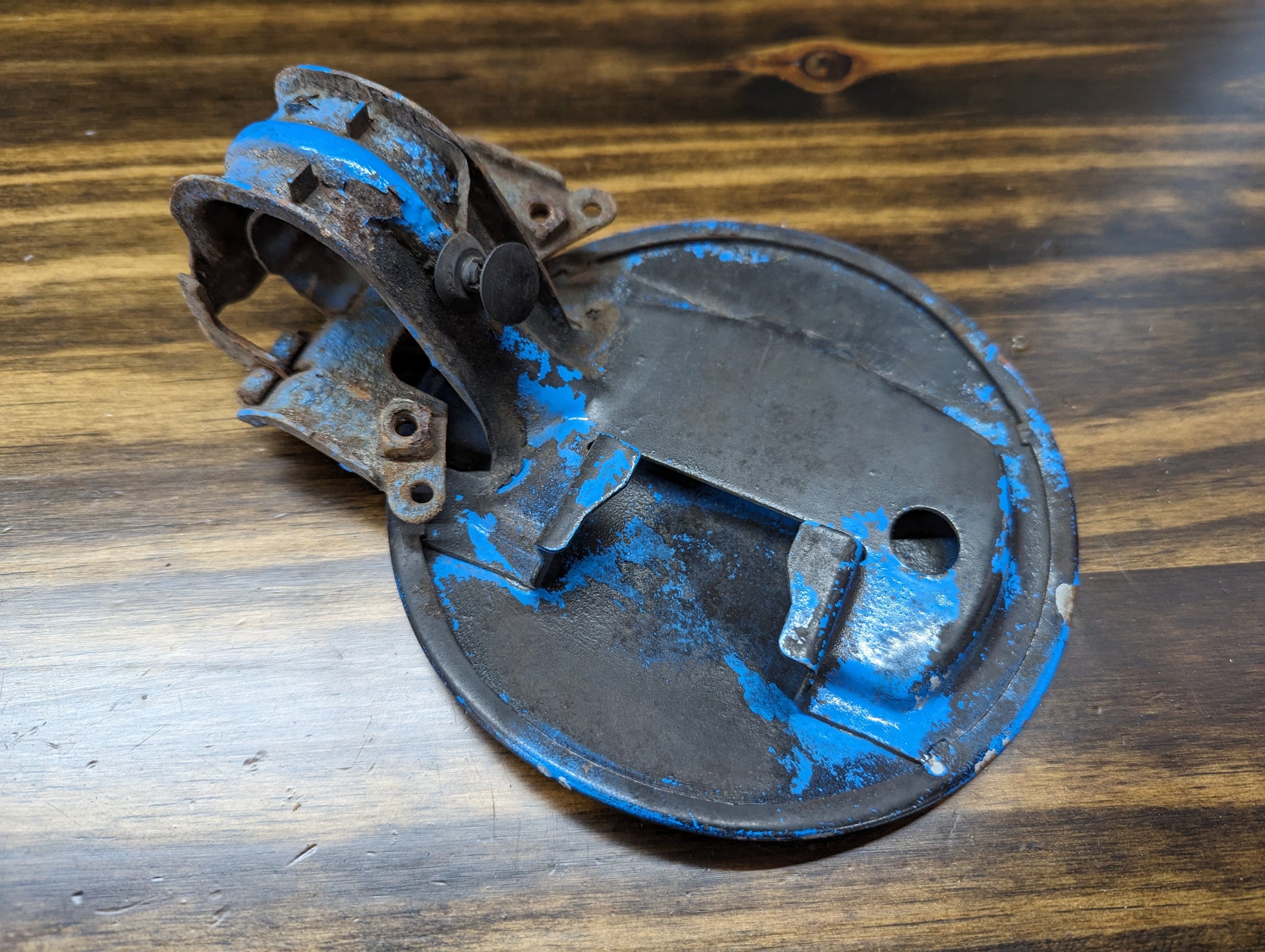A black OEM Fuel Filler Neck Door Gas Tank Cover by General Motors for 2001-2004 Chevy S10 & GMC Sonoma, featuring a hinge and latch mechanism. Paint wear visible with some bare metal exposed; item shown resting on wood surface.