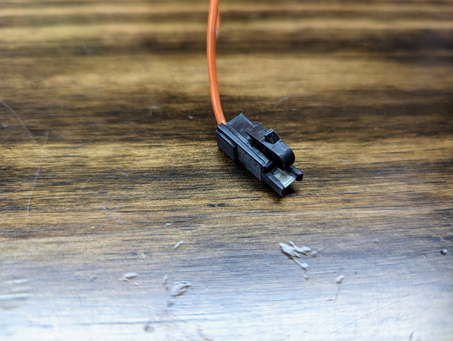A close-up of an Aptiv KIT Power Tap Connector & Terminals for Under Dash Fuse Panel, compatible with many 80s & 90s vehicles, featuring a black connector and orange wire on a scratched wooden surface.