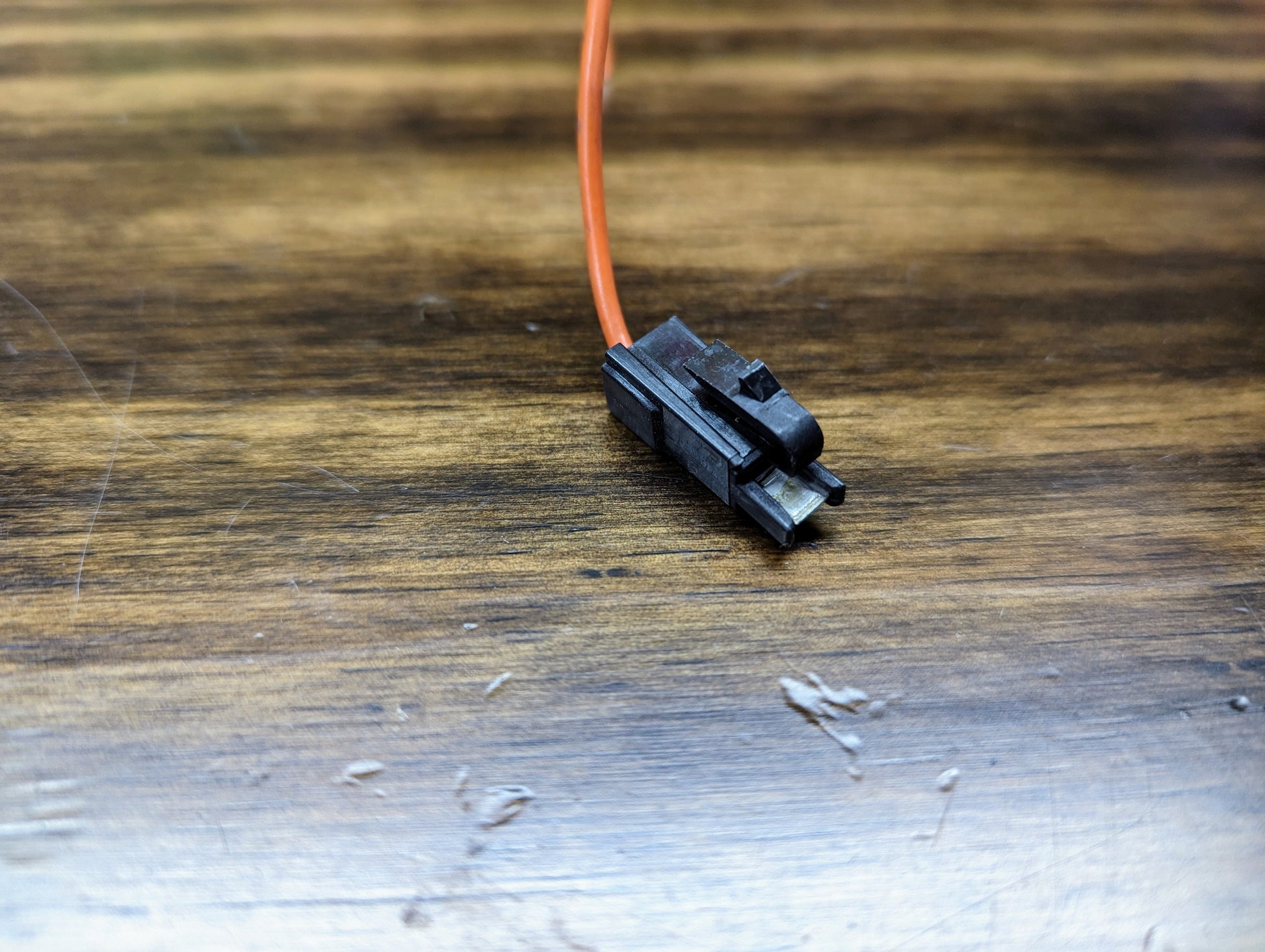 A close-up of an Aptiv KIT Power Tap Connector & Terminals for Under Dash Fuse Panel, compatible with many 80s & 90s vehicles, featuring a black connector and orange wire on a scratched wooden surface.