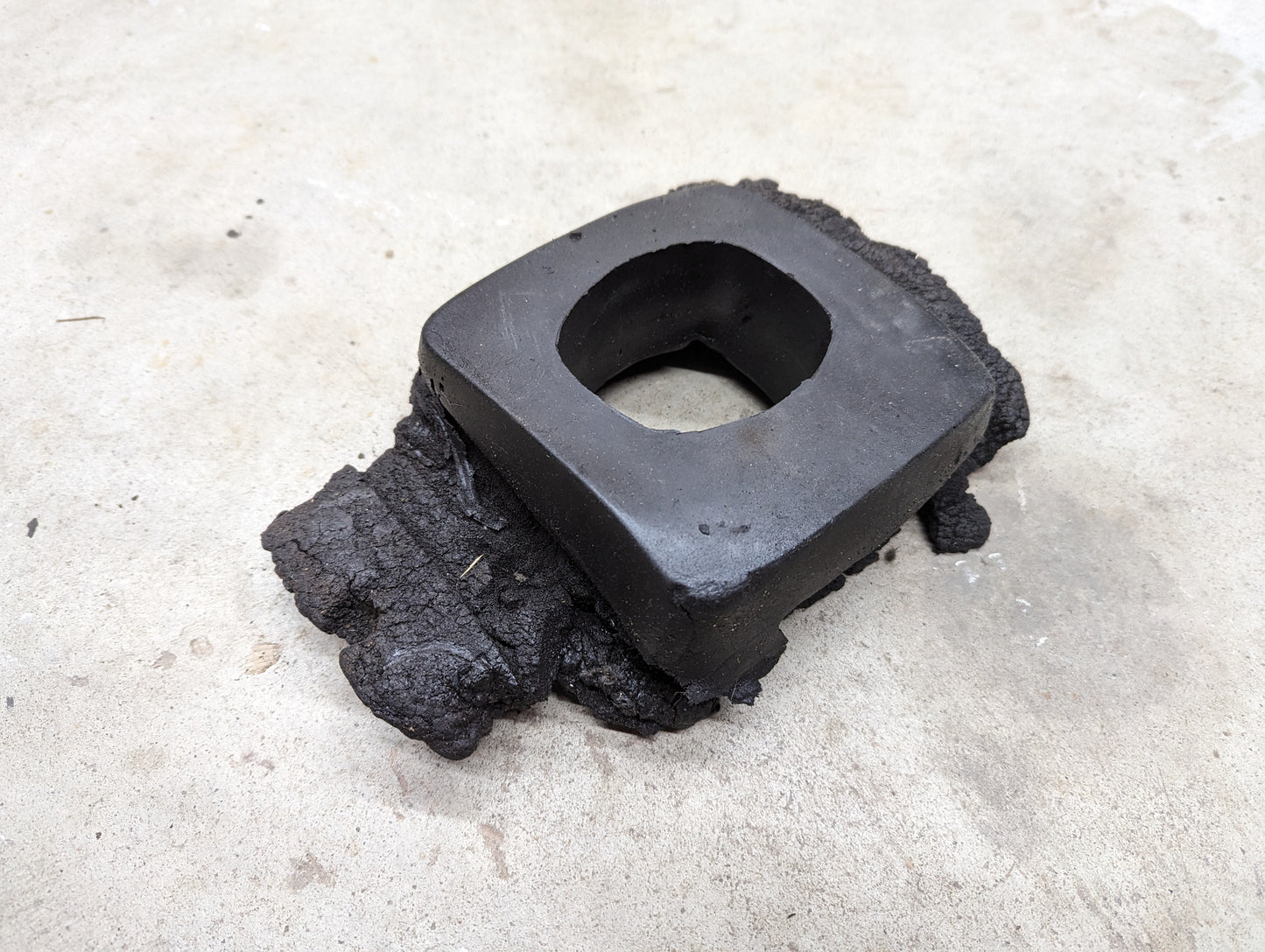 A black, rectangular OEM Manual Floor Shifter Insulator Foam by General Motors, designed for 1998-2005 Chevy S10 and Blazer, with a central square hole and rough fused material, rests on concrete.