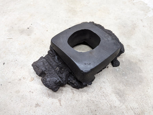 A black, rectangular OEM Manual Floor Shifter Insulator Foam by General Motors, designed for 1998-2005 Chevy S10 and Blazer, with a central square hole and rough fused material, rests on concrete.