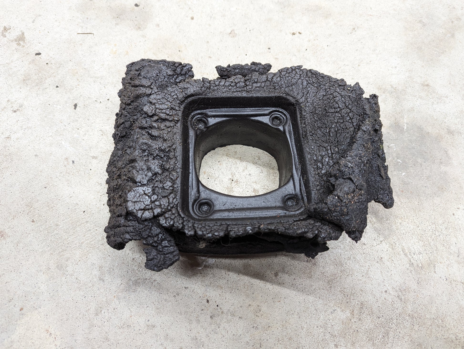 A black, rectangular General Motors OEM Manual Floor Shifter Insulator Foam for 1998–2005 Chevy S10 and Blazer with a hollow square center and four bolt holes rests on rough concrete, showing signs of damage.