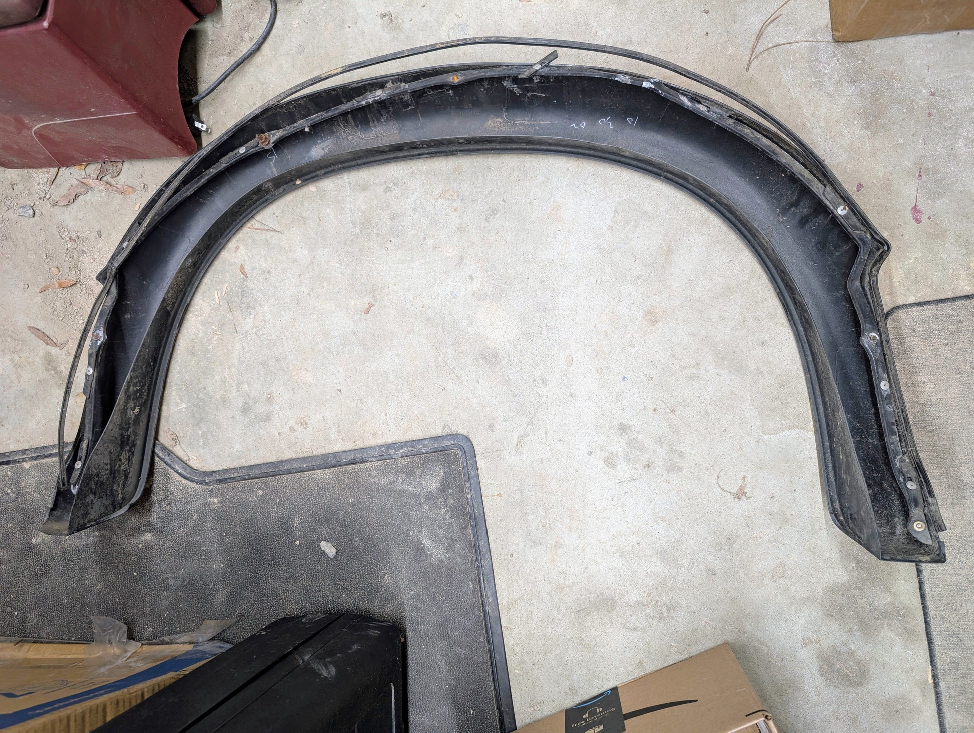 A General Motors OEM passenger rear bed flare extension for a 1994-2004 Chevy S10 or GMC Sonoma rests on a concrete floor, partly on a black mat, with cardboard boxes, a maroon item, and scattered debris nearby.
