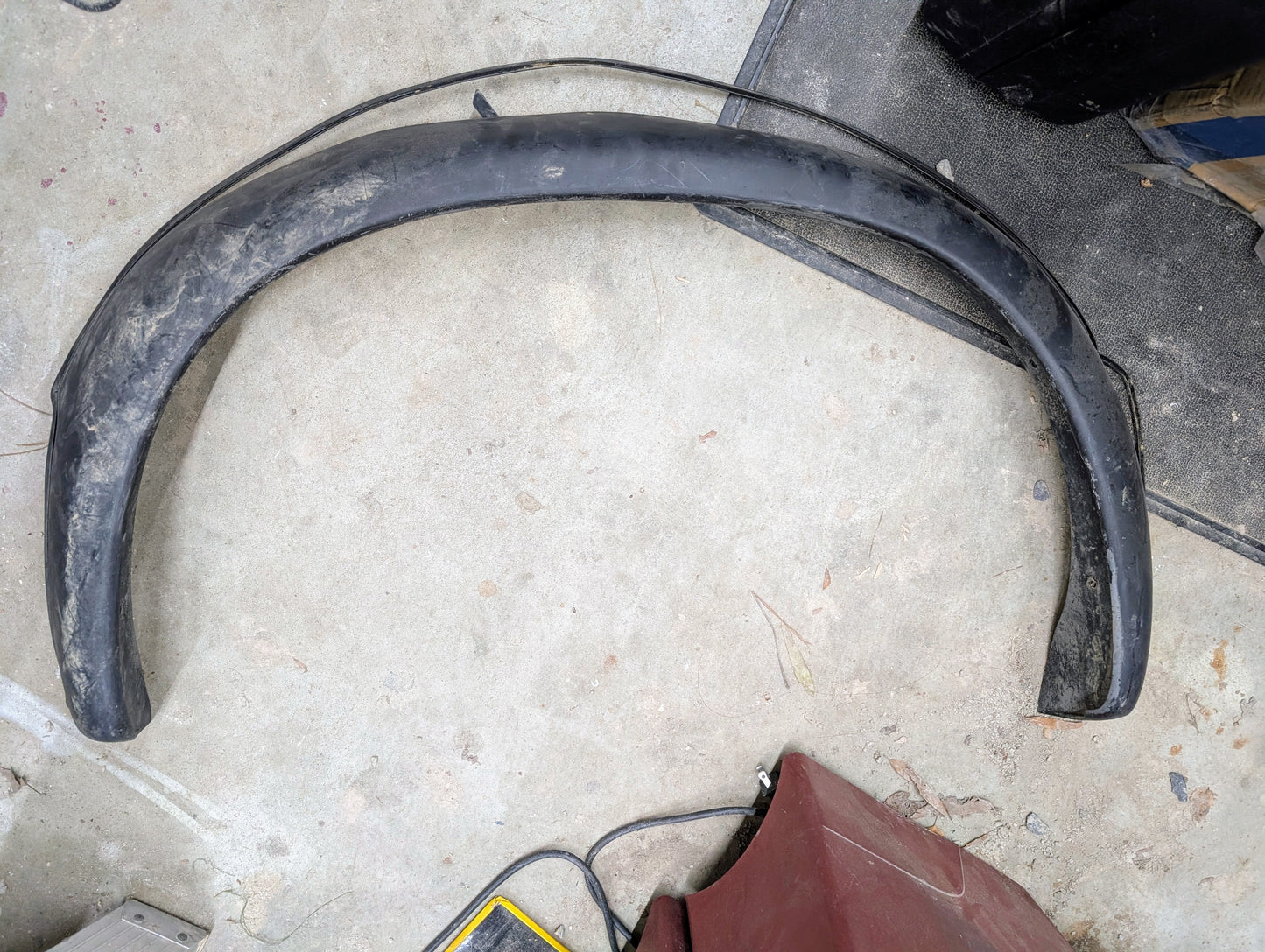 A General Motors OEM passenger rear bed flare extension for a 1994-2004 Chevy S10 or GMC Sonoma rests on a dirty, scratched concrete surface, with parts of a red object and an item with yellow edges nearby.