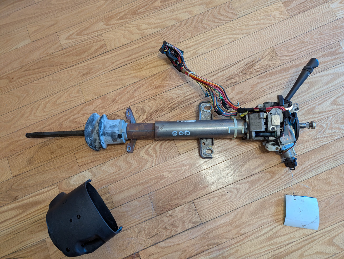 A disassembled General Motors OEM Steering Column w/Tilt, Cruise Control, and Console Shift for 1998-2005 Chevy S10 Blazer and more lies on a wooden floor with wiring, control levers, black plastic cover, and a small metal plate nearby.