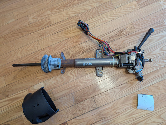 A disassembled General Motors OEM Steering Column w/Tilt, Cruise Control, and Console Shift for 1998-2005 Chevy S10 Blazer and more lies on a wooden floor with wiring, control levers, black plastic cover, and a small metal plate nearby.