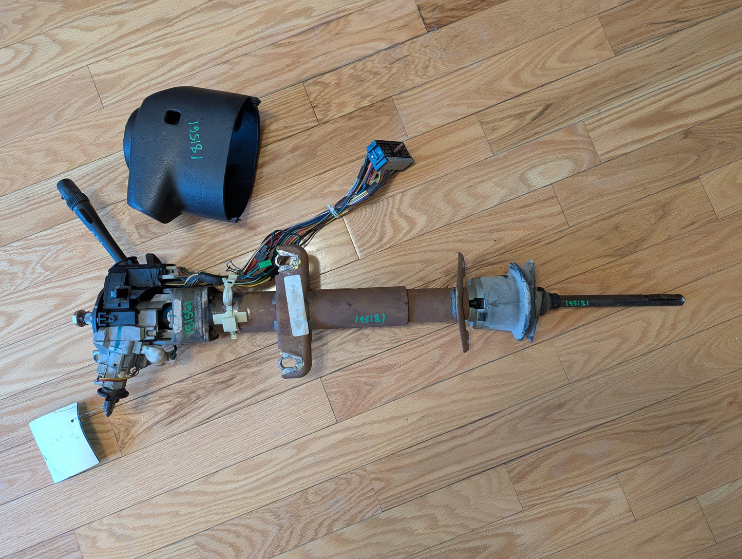 A disassembled General Motors OEM Steering Column with tilt, cruise control, and console shift for 1998-2005 Chevy S10 Blazer rests on a light wood floor, showing metal parts, wiring, connectors, and a black plastic cover.