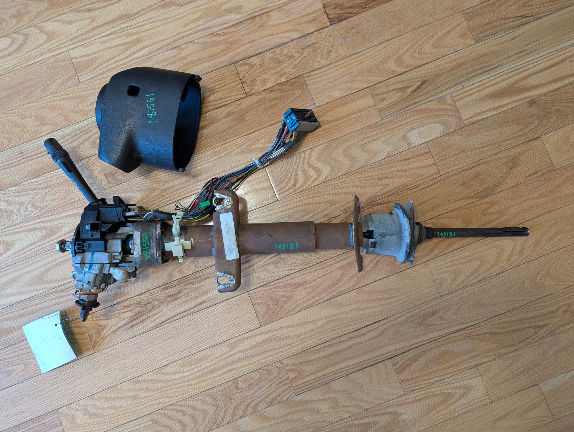 A disassembled General Motors OEM Steering Column with tilt, cruise control, and console shift for 1998-2005 Chevy S10 Blazer rests on a light wood floor, showing metal parts, wiring, connectors, and a black plastic cover.