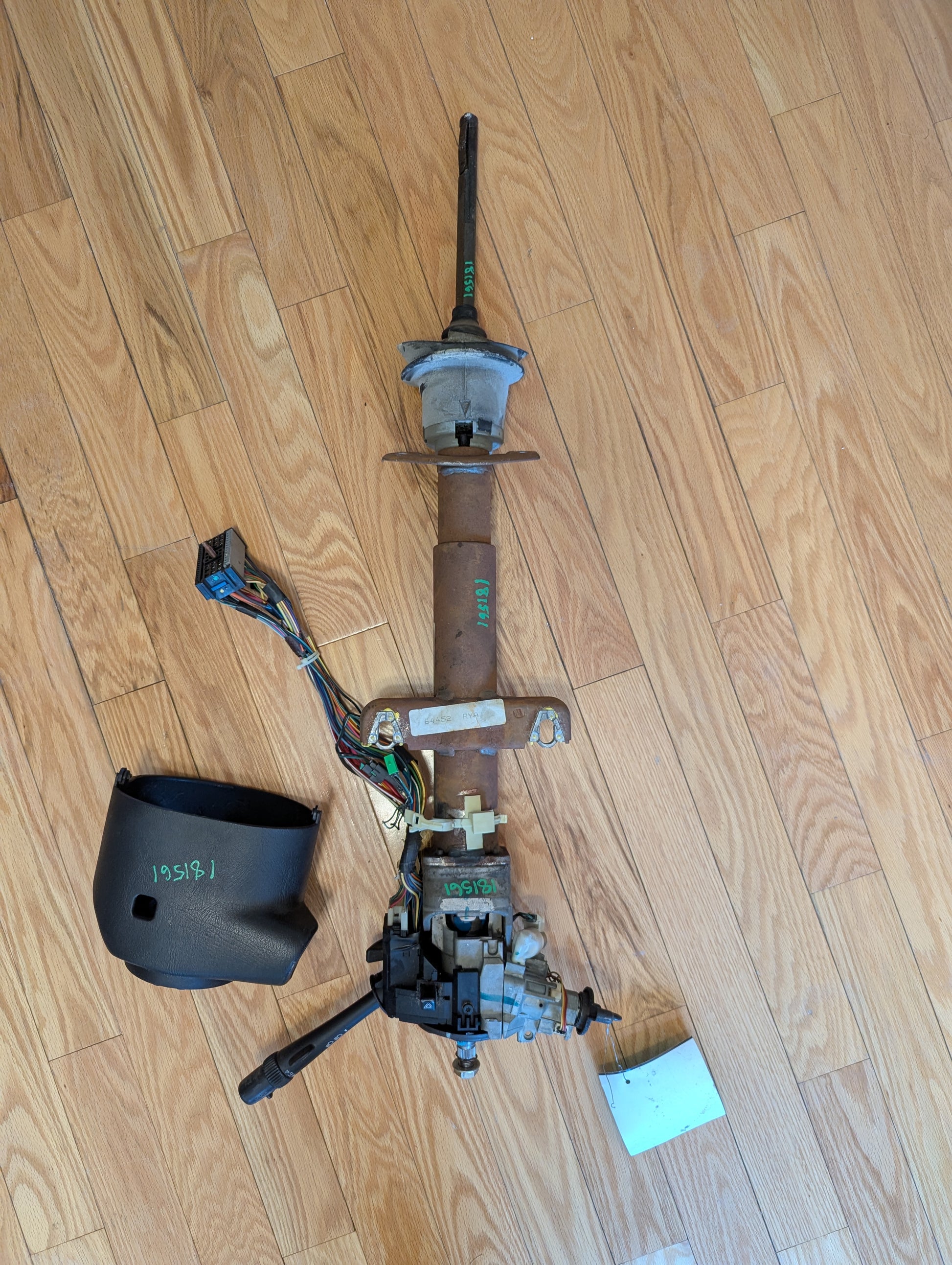 A disassembled General Motors OEM Steering Column w/Tilt, Cruise Control, and Console Shift for 1998-2005 Chevy S10 Blazer is shown on a wood floor with its black plastic cover, attached wires, levers, and a small white paper.