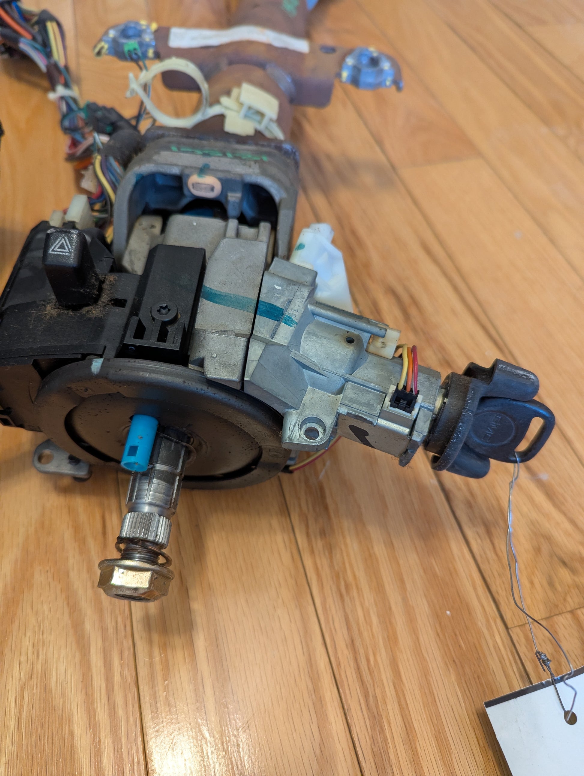 A close-up of a General Motors OEM Steering Column with Tilt, Cruise Control, and Console Shift for 1998-2005 Chevy S10 Blazer, showing wiring and car key inserted, placed on a wooden floor.