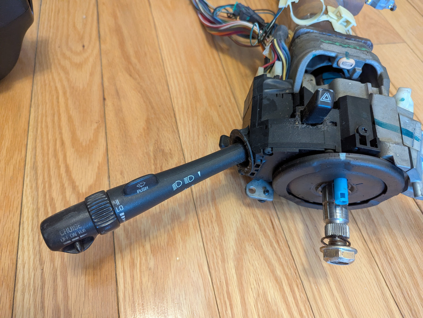 A General Motors OEM steering column with tilt, cruise control, and console shift for 1998-2005 Chevy S10 Blazer and more is shown on a wooden floor, highlighting the lever with light and wiper markings.