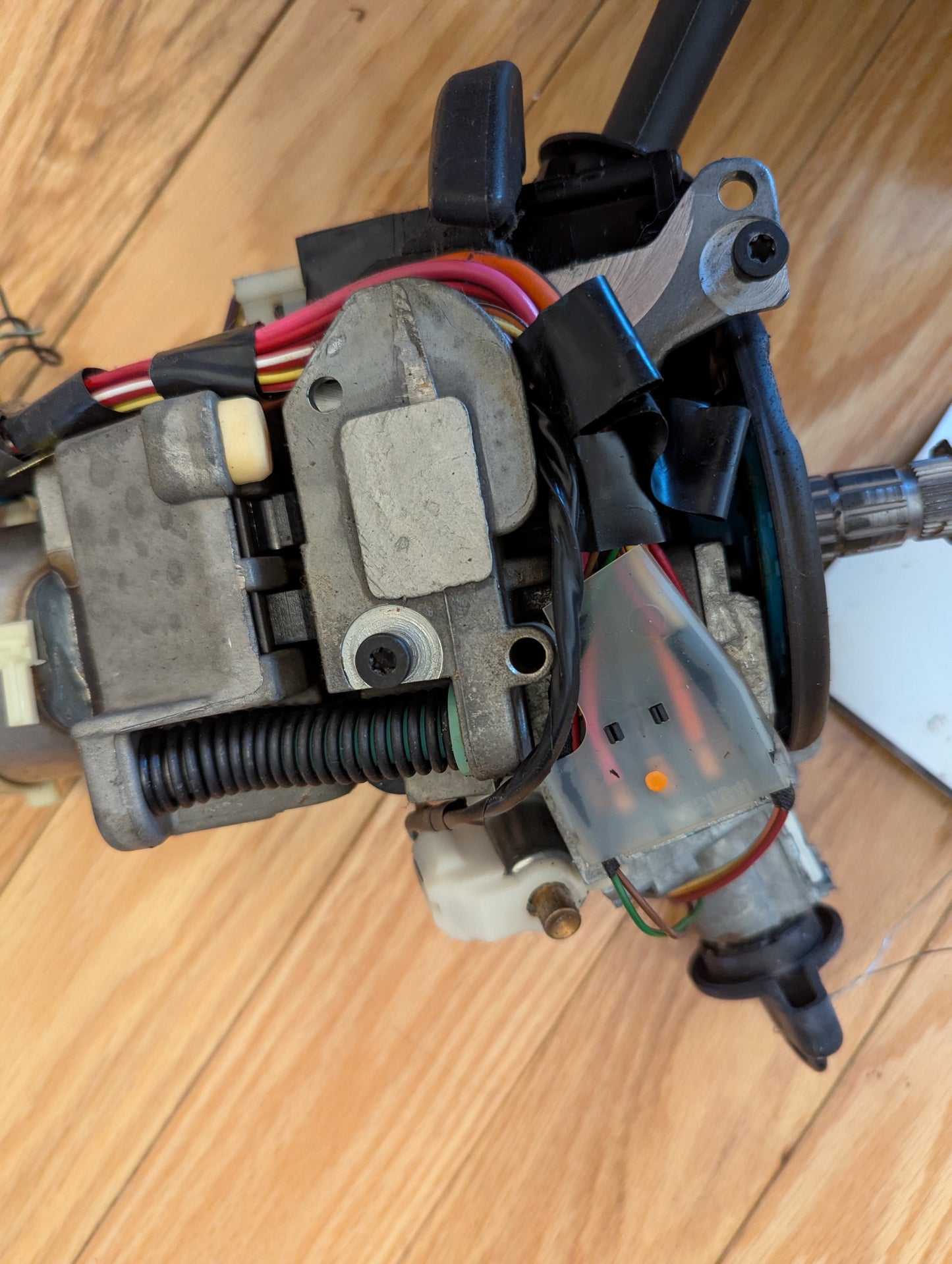Close-up of an OEM Steering Column with tilt, cruise control, and console shift for 1998-2005 Chevy S10 Blazer by General Motors, showing metal parts, colored wires, and a clear plastic connector with orange markings on a wooden surface.