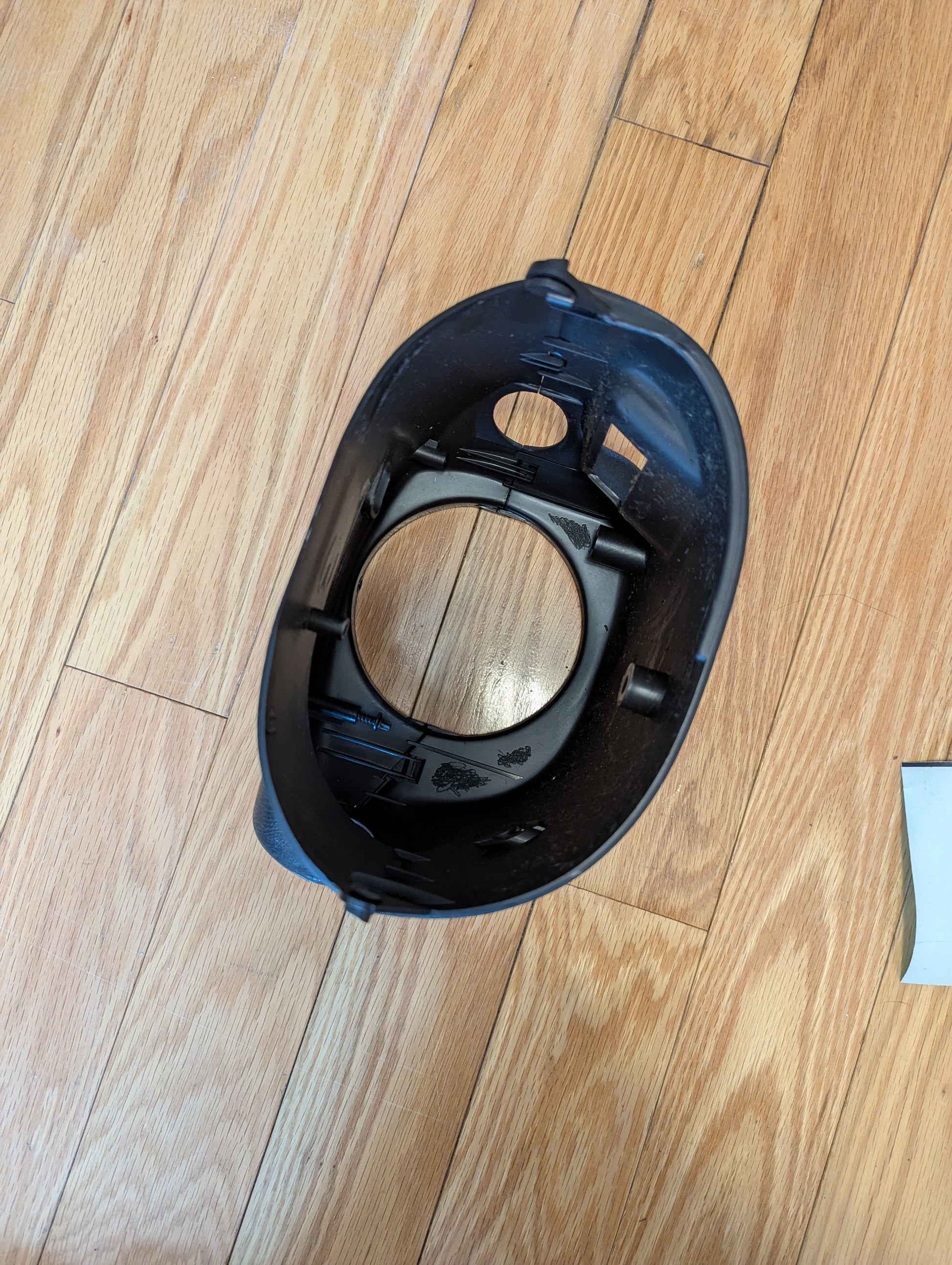 A black plastic part from a General Motors OEM Steering Column w/Tilt, Cruise Control, and Console Shift for 1998-2005 Chevy S10 Blazer rests on a light wood floor, oval-shaped with clips and a large central hole visible.