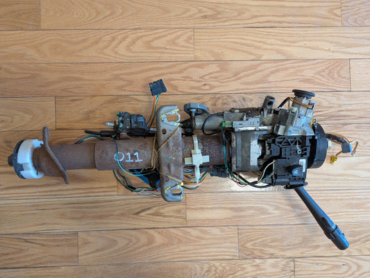 A used General Motors OEM Steering Column for 2003-2006 GM Full Size Trucks, disassembled with exposed wires, connectors, brackets, and a black lever, is laid out on a wooden floor.