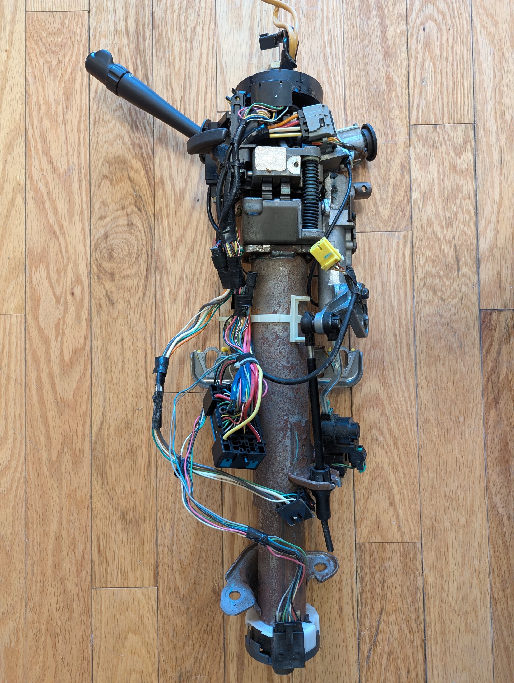 A General Motors OEM Steering Column for 2003-2006 GM Full Size Trucks, featuring exposed wiring, switches, and connectors, rests on a wood floor. Colored wires and components are visible on the metal automatic transmission column.