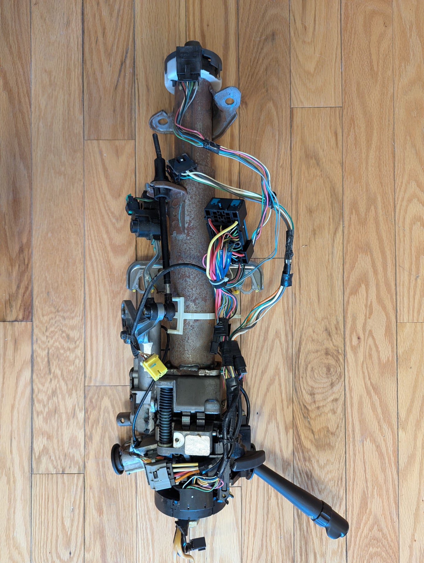 An OEM Steering Column for 2003-2006 GM Full Size Trucks by General Motors, showing exposed wires, connectors, and mechanical parts, is displayed disassembled on a light wood floor.
