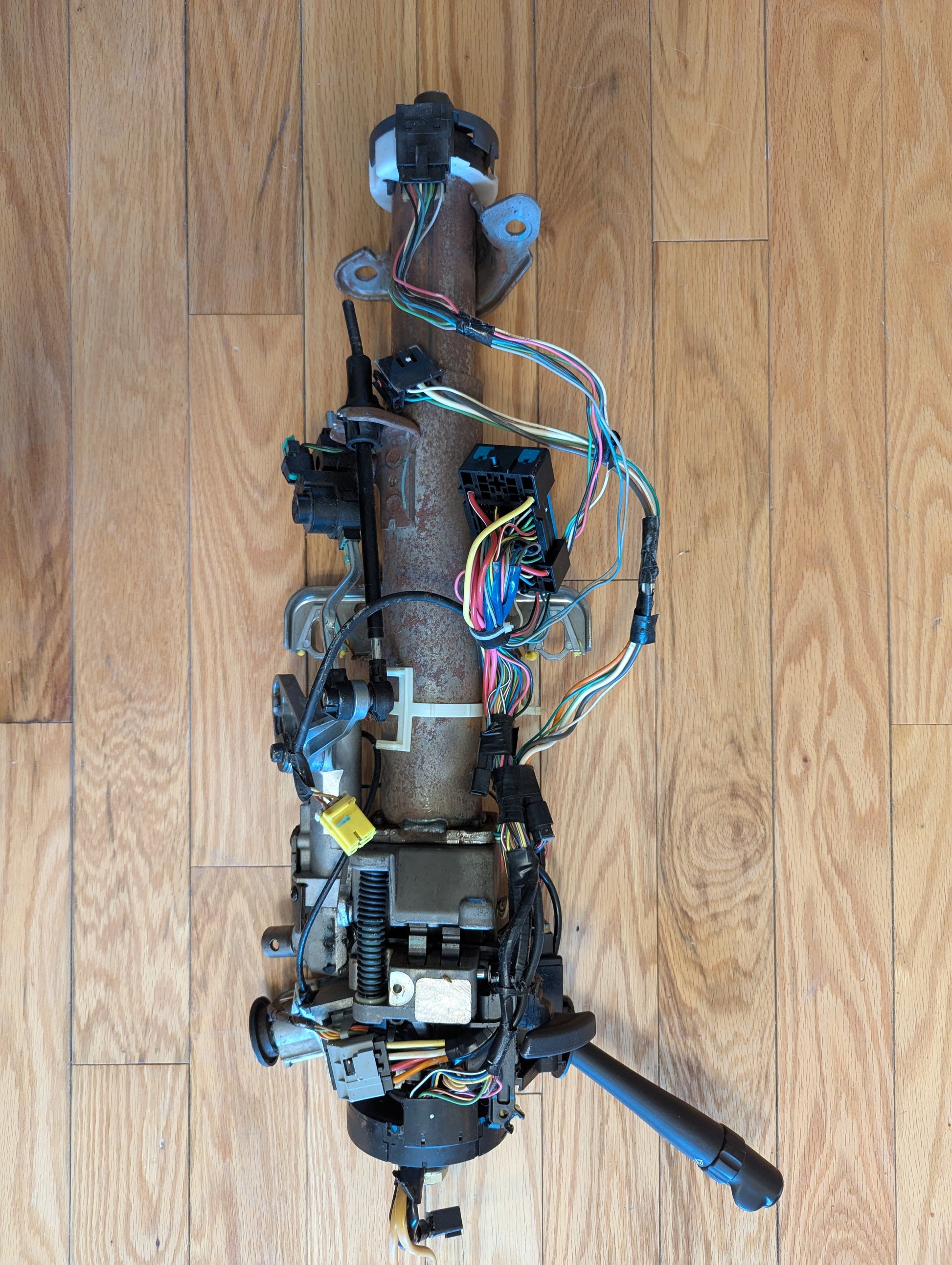 An OEM Steering Column for 2003-2006 GM Full Size Trucks by General Motors, showing exposed wires, connectors, and mechanical parts, is displayed disassembled on a light wood floor.