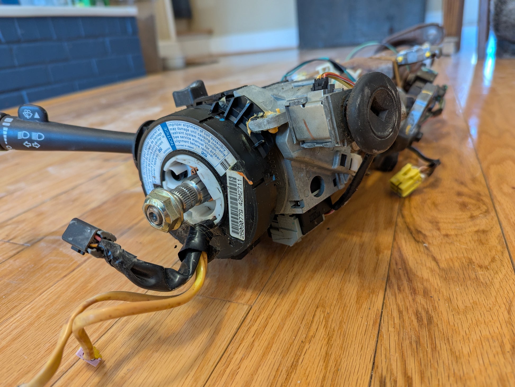 An OEM Steering Column for 2003-2006 GM Full Size Trucks by General Motors, shown disassembled with wiring and switches exposed, rests on a wooden floor indoors.