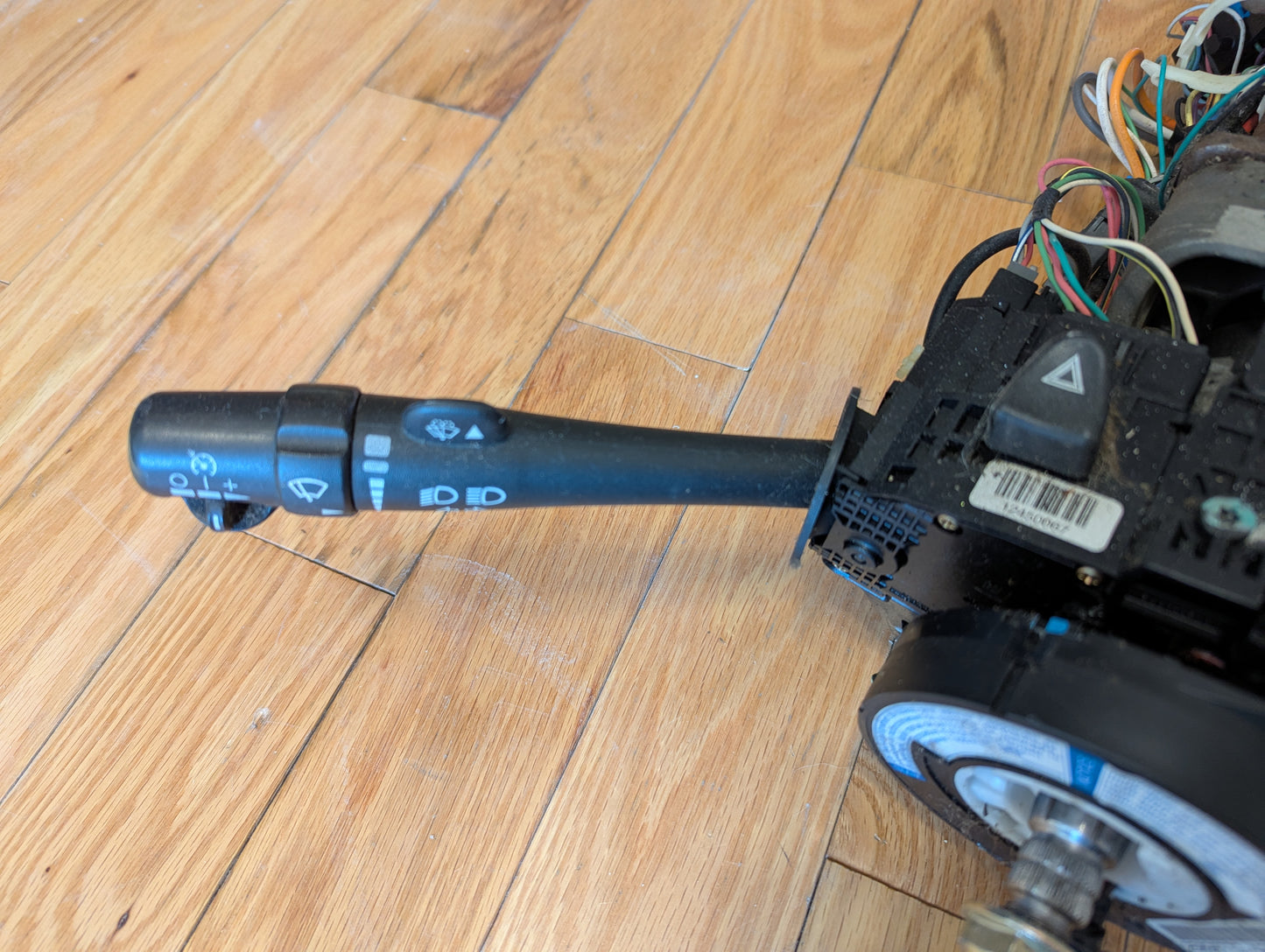 A close-up of a General Motors OEM Steering Column for 2003-2006 GM Full Size Trucks shows the turn signal and windshield wiper stalks with exposed wires, all attached to part of the assembly and placed on a wooden floor.