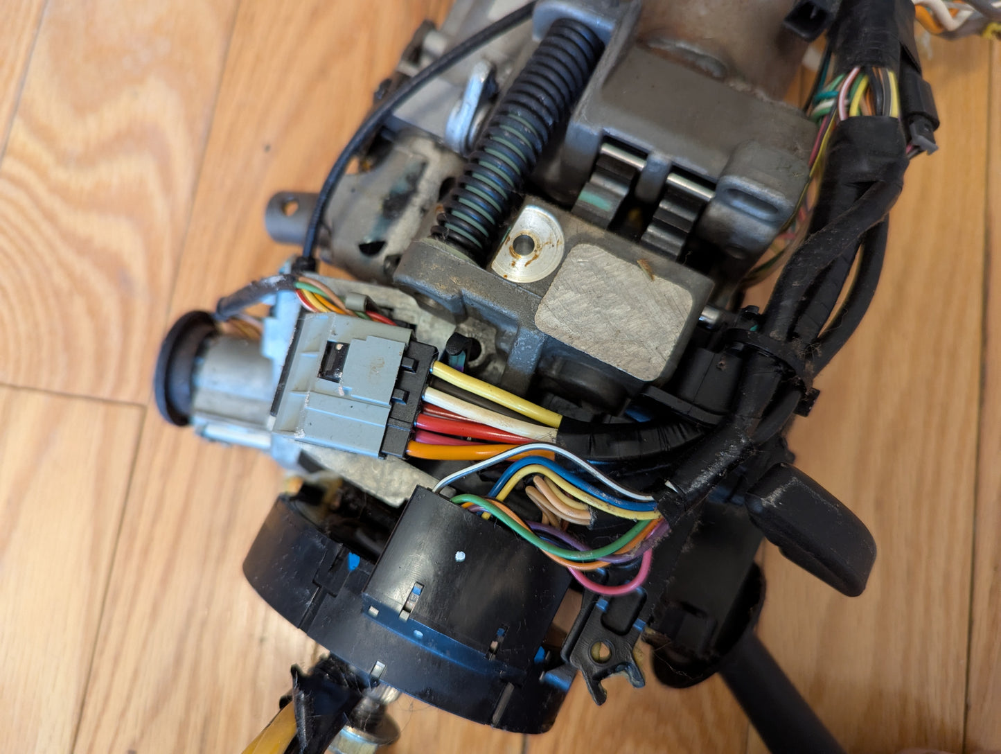 A close-up of a General Motors OEM Steering Column for 2003-2006 GM Full Size Trucks, showing colored wires, connectors, and mechanical parts on a wooden surface.