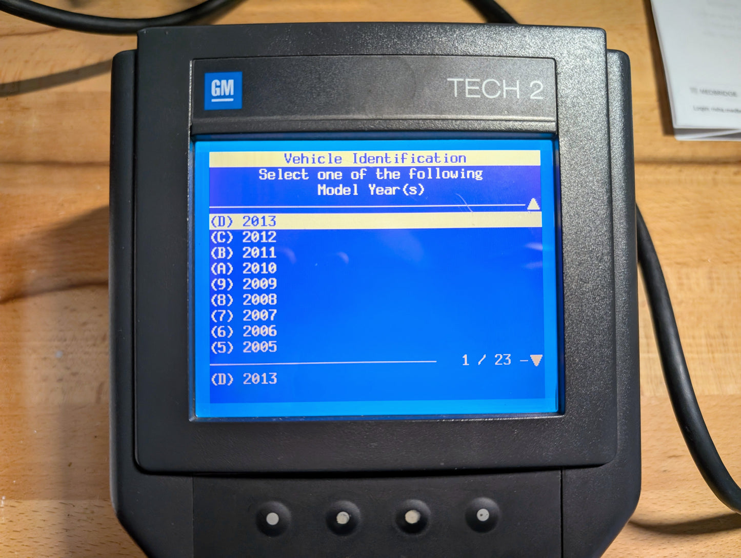 A Vetronix TESTED Tech 2 Automotive Diagnostic Tool & Accessories for 1991-2013 GM Vehicles is shown on a wooden surface, displaying a blue screen with selectable model years (2013 to 2006) and visible cables.