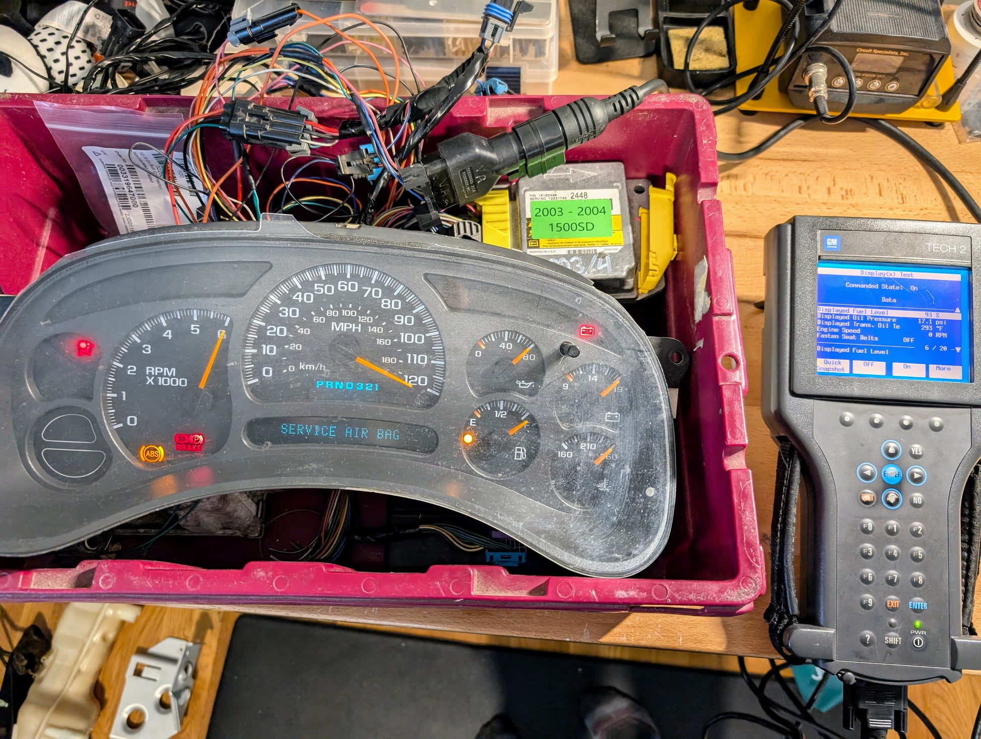 A Vetronix TESTED Tech 2 Automotive Diagnostic Tool & Accessories for 1991-2013 GM vehicles is connected to an instrument cluster with warning lights and wiring in a red bin, next to a workbench displaying vehicle data.