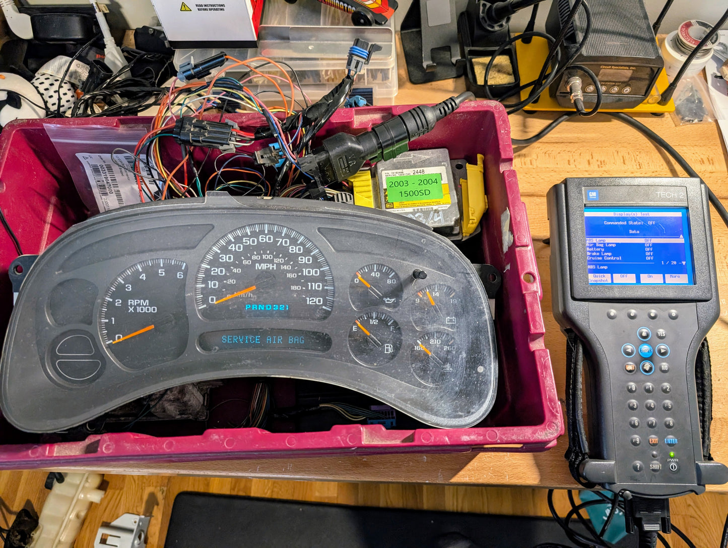 A car instrument cluster rests in a pink bin with various wires and a Vetronix TESTED Tech 2 Automotive Diagnostic Tool & Accessories for 1991-2013 GM Vehicles, displayed on a workbench with electronics in the background.