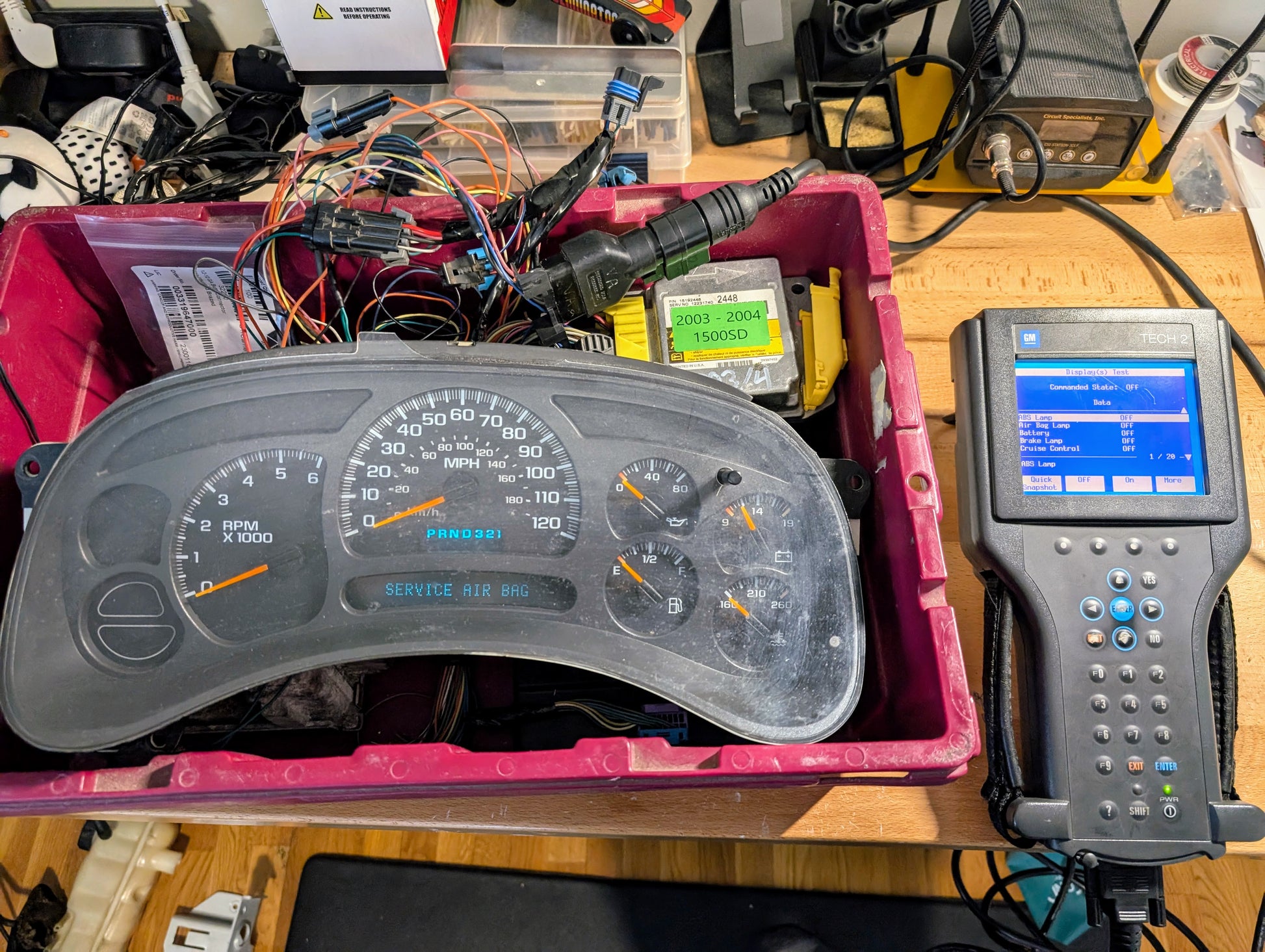 A car instrument cluster rests in a pink bin with various wires and a Vetronix TESTED Tech 2 Automotive Diagnostic Tool & Accessories for 1991-2013 GM Vehicles, displayed on a workbench with electronics in the background.