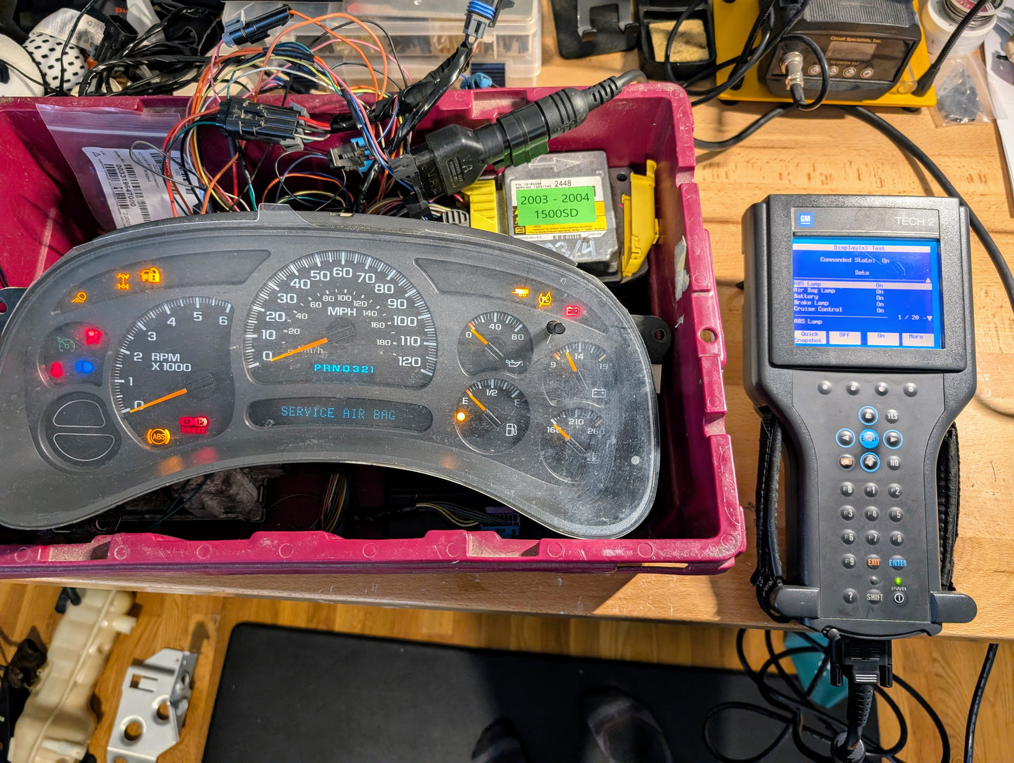 A Vetronix TESTED Tech 2 Automotive Diagnostic Tool & Accessories for 1991-2013 GM Vehicles is connected to a powered-on car instrument cluster, with wires and components arranged in a red plastic tray on the workbench.