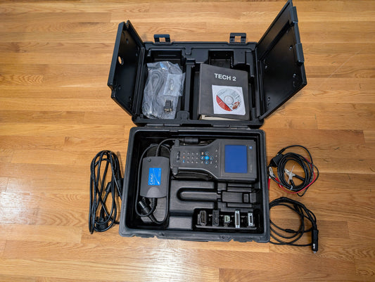 An open black case on a wooden floor holds a Vetronix TESTED Tech 2 Automotive Diagnostic Tool & Accessories for 1991-2013 GM vehicles, including cables, adapters, handheld device, manuals, and software CD.