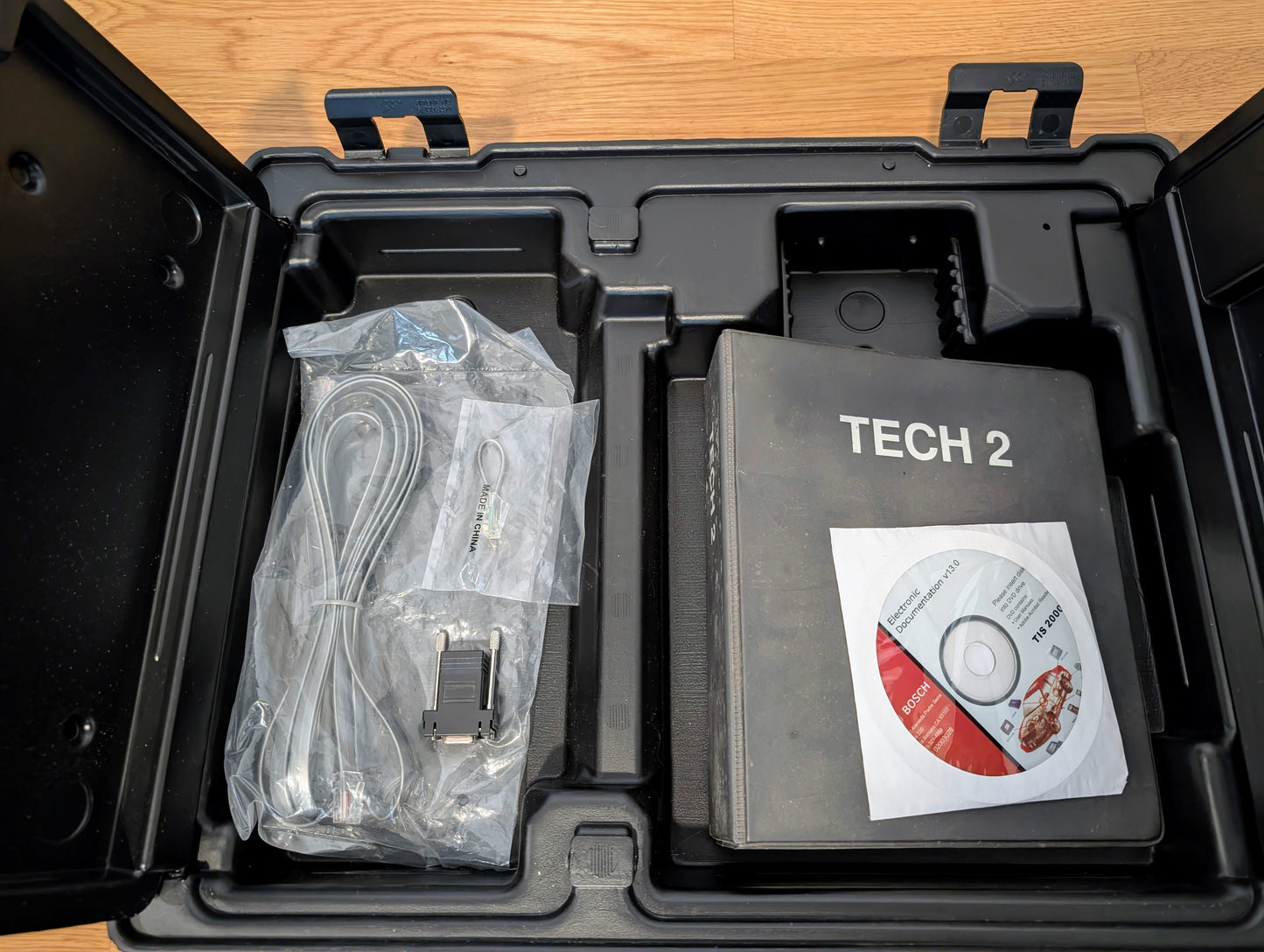 An open black plastic Vetronix case on a wooden floor holds a TESTED Tech 2 Automotive Diagnostic Tool & Accessories for 1991-2013 GM vehicles, including various cables and a Tech 2 Software CD, all neatly arranged in molded compartments.