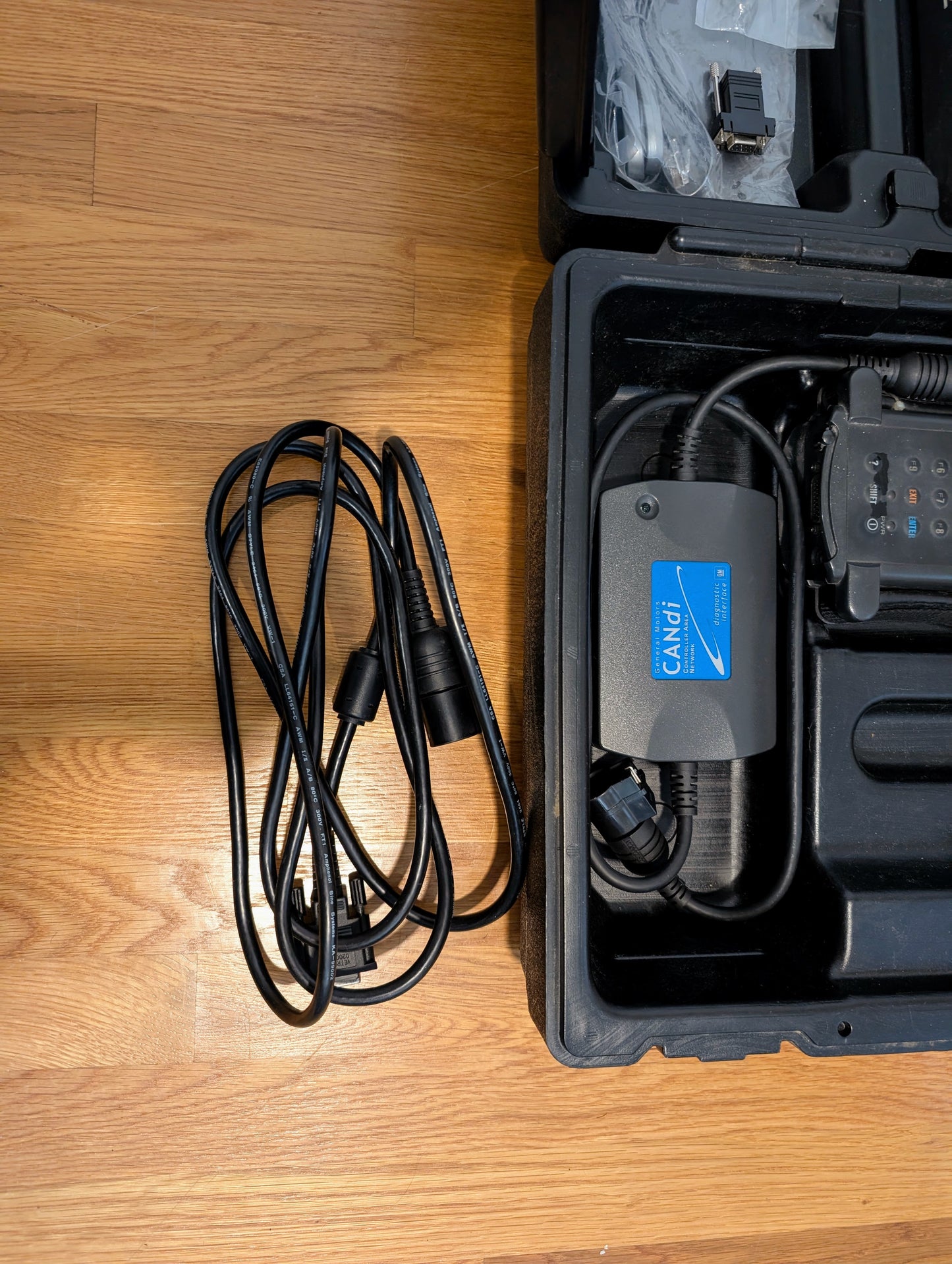 A black charging cable lies coiled on a wooden floor beside an open black case holding a Vetronix TESTED Tech 2 Automotive Diagnostic Tool & Accessories for 1991-2013 GM Vehicles, including the CANdi module and more.