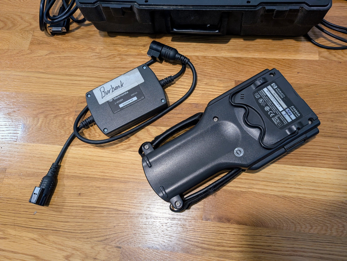 A Vetronix TESTED Tech 2 Automotive Diagnostic Tool & accessories for 1991-2013 GM vehicles, along with a Burbank power adapter, are on a wooden floor; part of a black case is also visible in the background.
