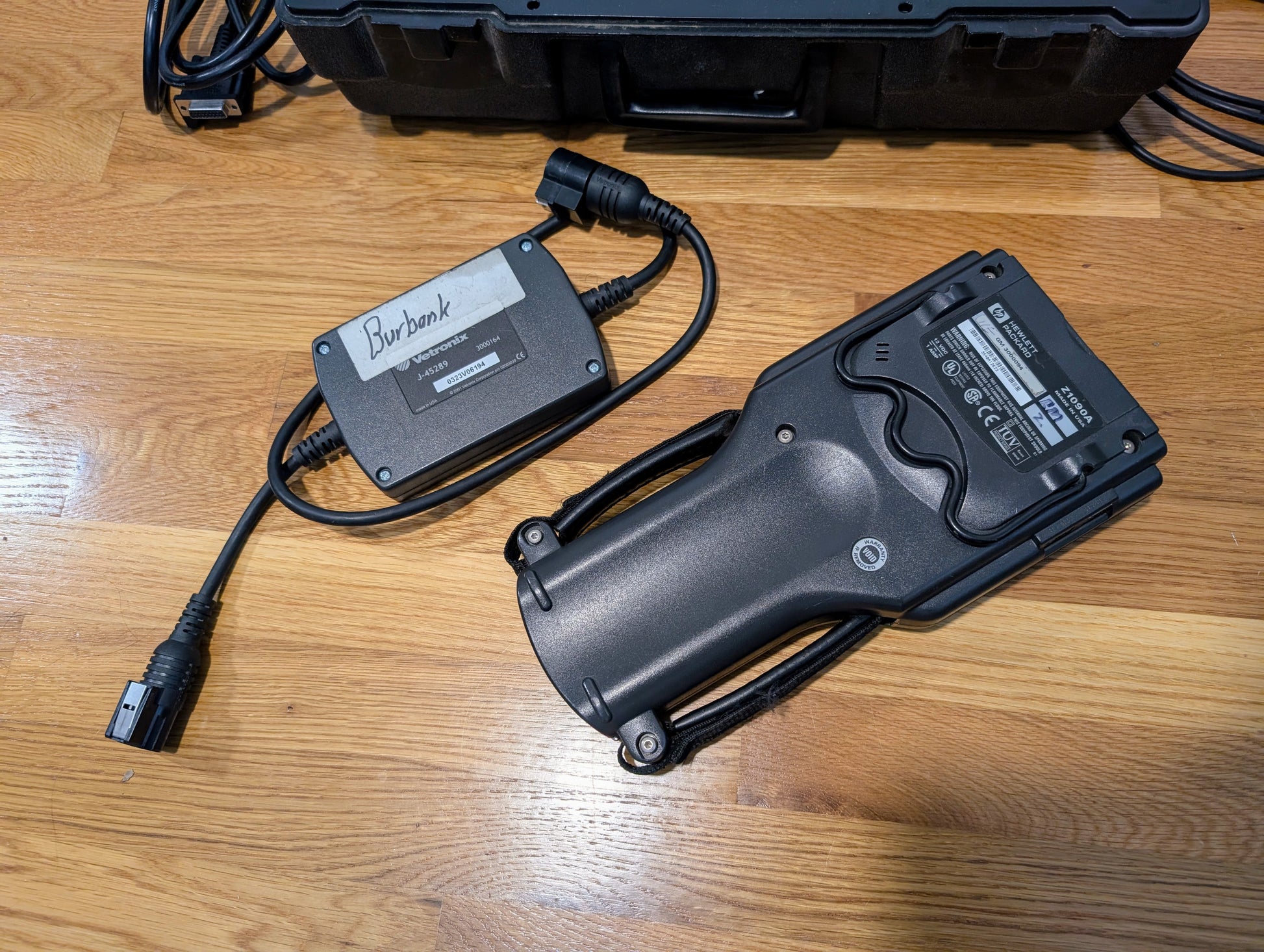 A Vetronix TESTED Tech 2 Automotive Diagnostic Tool & accessories for 1991-2013 GM vehicles, along with a Burbank power adapter, are on a wooden floor; part of a black case is also visible in the background.