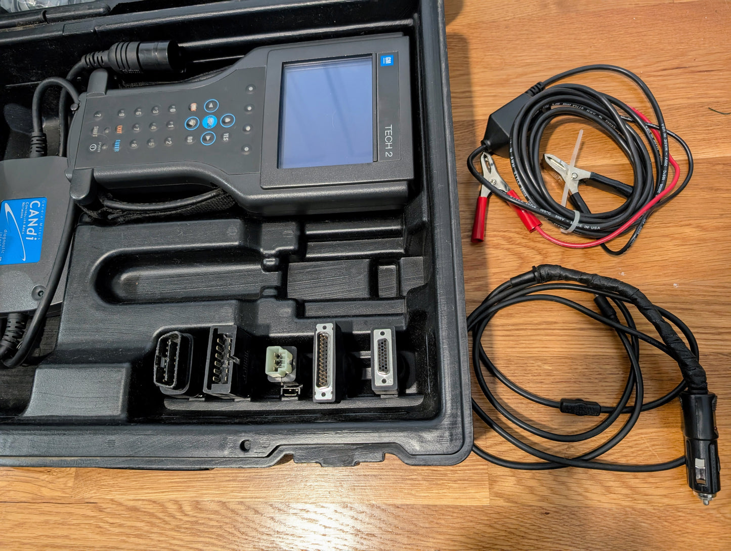A Vetronix TESTED Tech 2 Automotive Diagnostic Tool & Accessories for 1991-2013 GM Vehicles, including connectors and cables like alligator clips and an OBD2 Code Scanner, is displayed in its carrying case on a wooden floor.