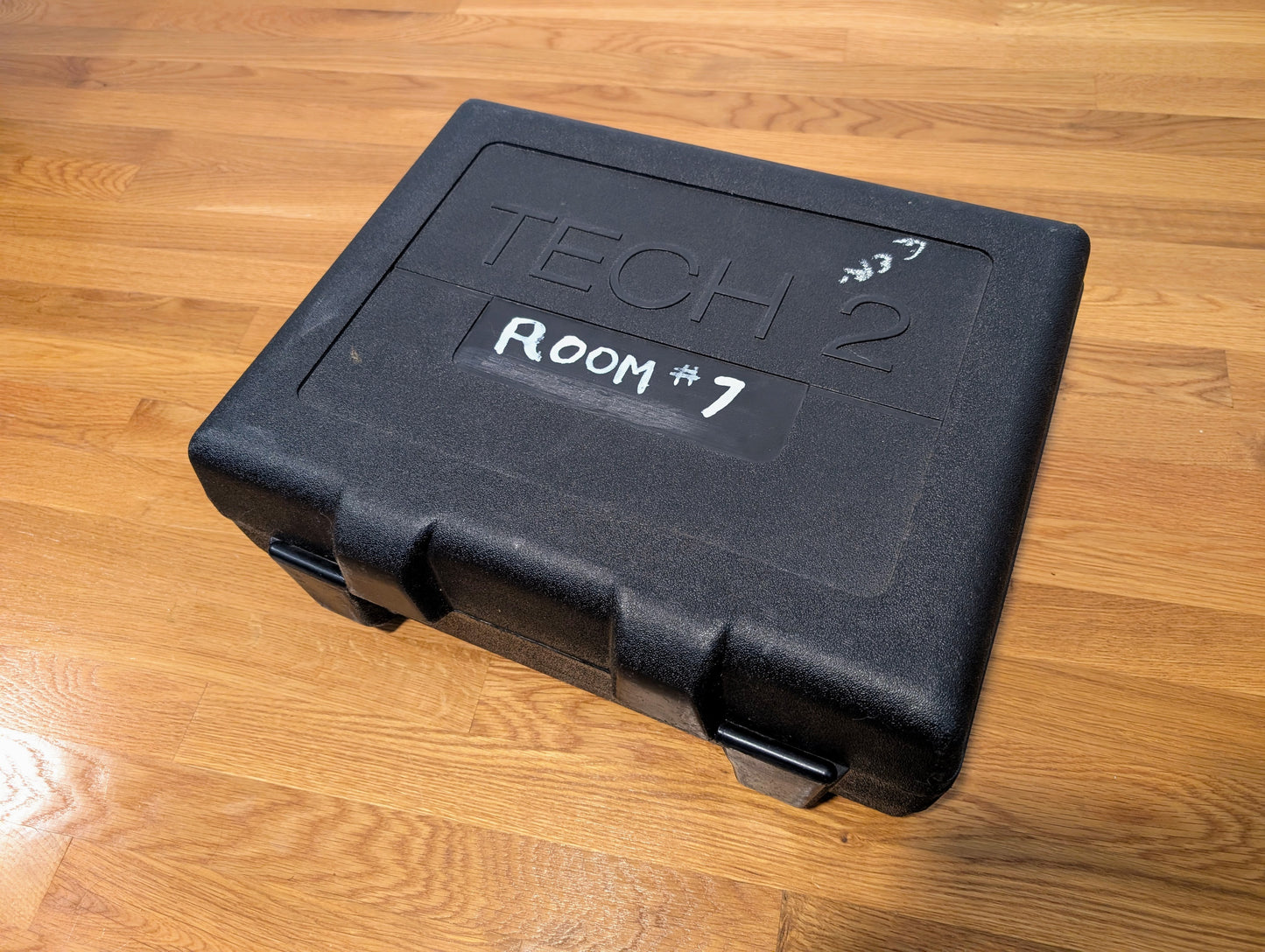 A black hard plastic Vetronix case labeled TECH 2 with a white Room #7 sticker sits on a light wood floor—ideal for storing your TESTED Tech 2 Automotive Diagnostic Tool & Accessories for 1991-2013 GM Vehicles.