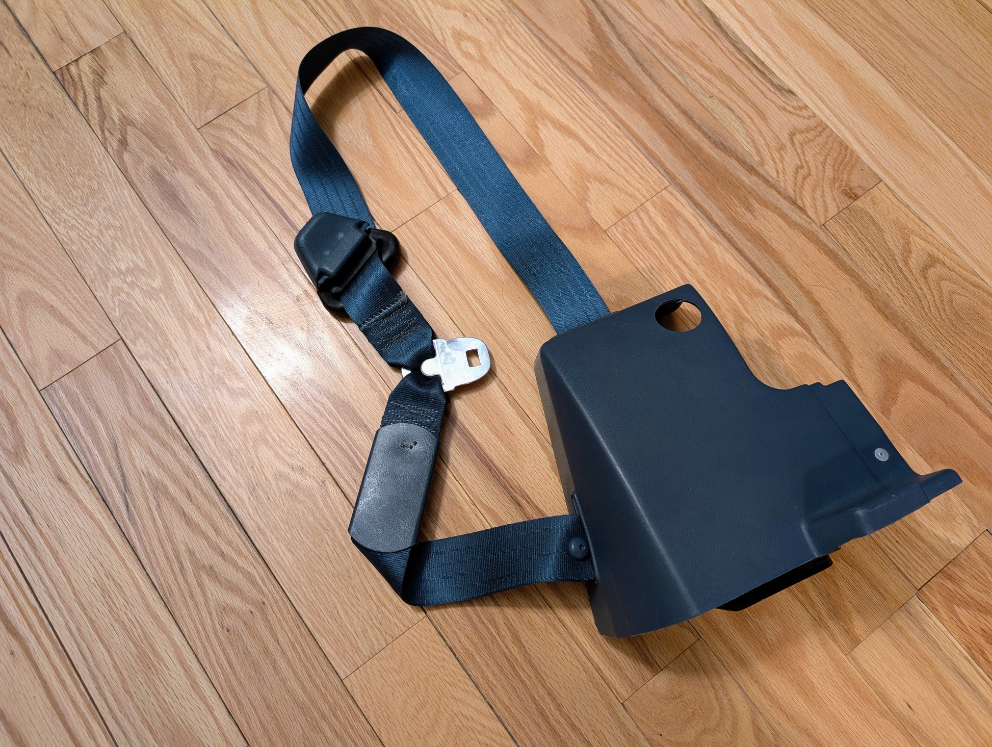 A General Motors NOS blue passenger seat belt retractor for 1982–1993 Chevy S10 and GMC S15 is shown with a plastic cover and latch laid out on a wooden floor.