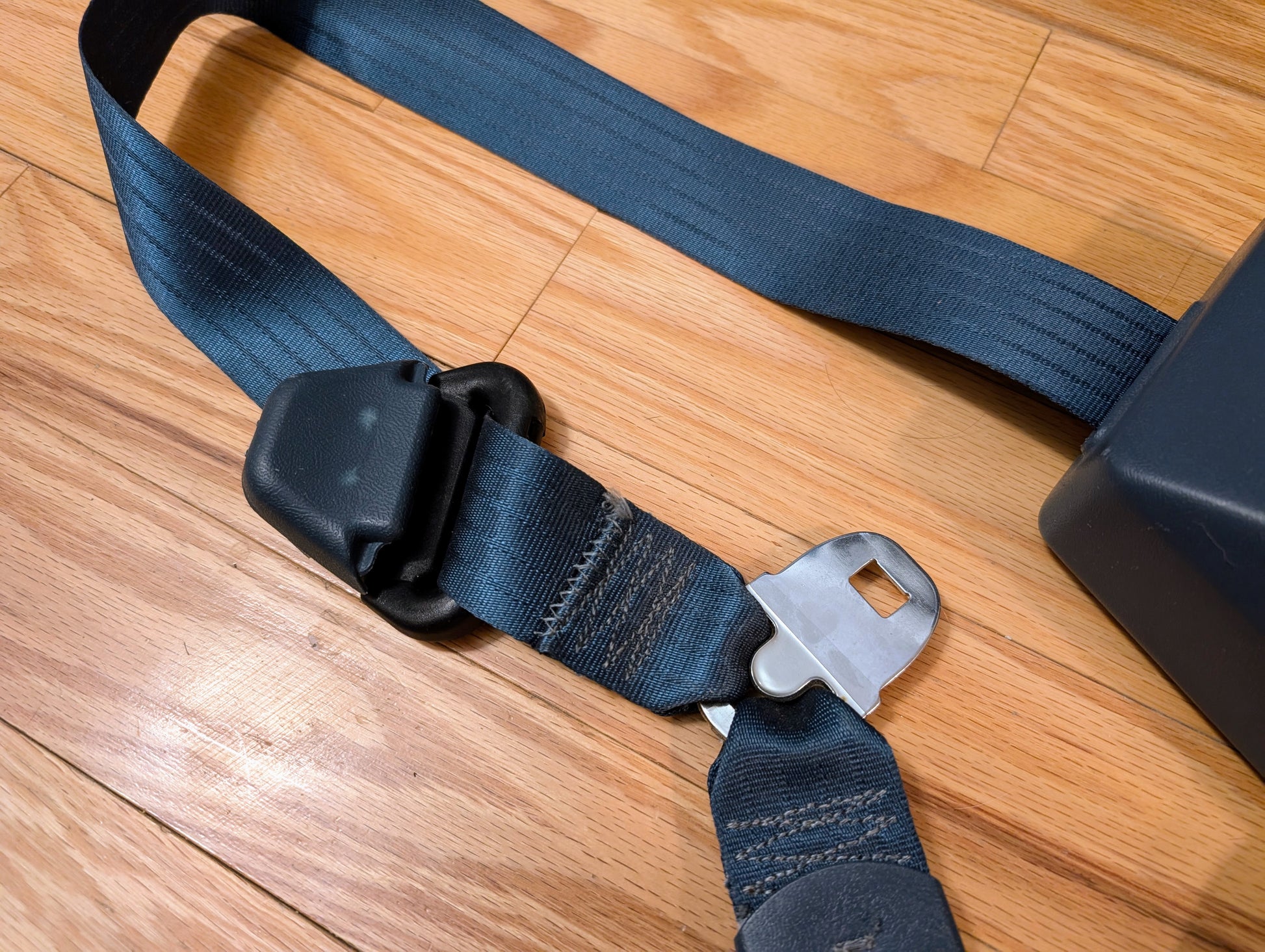 A close-up of a General Motors NOS Passenger Seat Belt Retractor in blue for 1982–1993 Chevy S10 and GMC S15, showing the buckle and metal latch on a wooden floor.