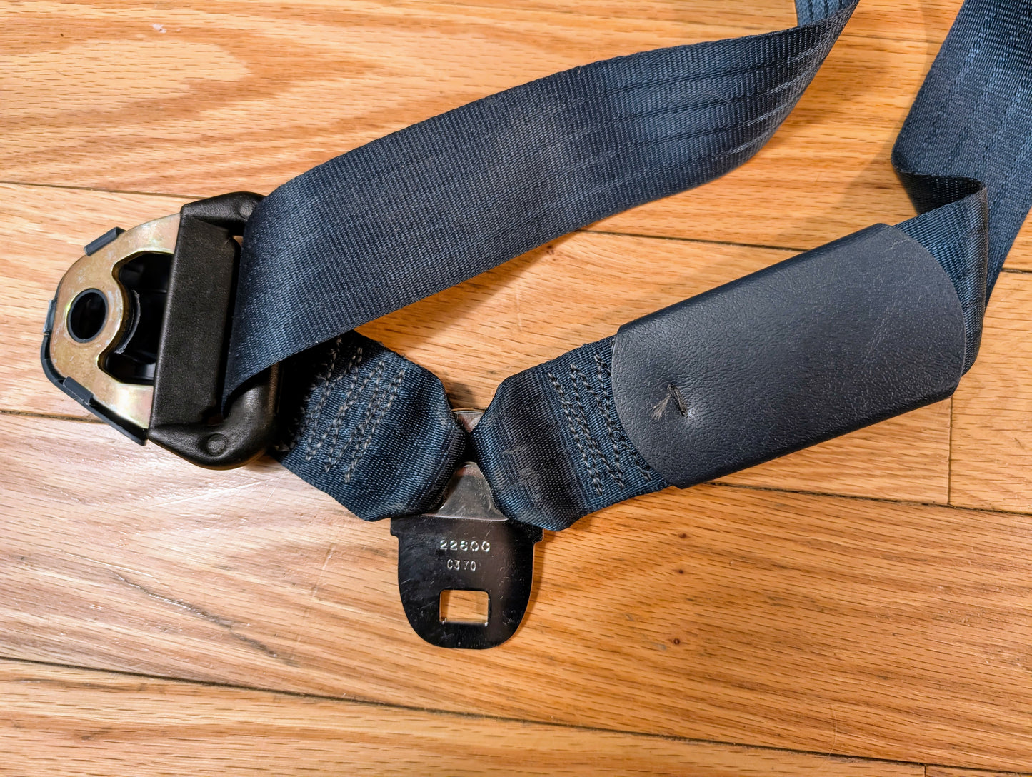A close-up of a General Motors NOS blue passenger seat belt retractor for 1982-1993 Chevy S10 or GMC S15 on a wooden floor, showing the buckle, latch plate, webbing, and stitching details.