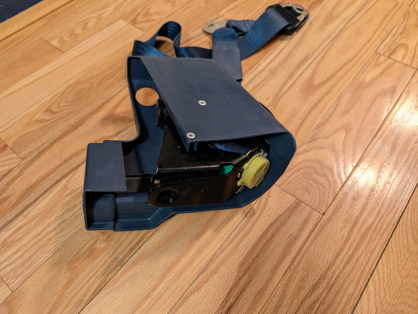 A NOS blue passenger seat belt retractor by General Motors for 1982-1993 Chevy S10 and GMC S15, featuring a metal buckle, plastic cover, and yellow button, lies on a wooden floor—ideal for OEM seat belt replacements.