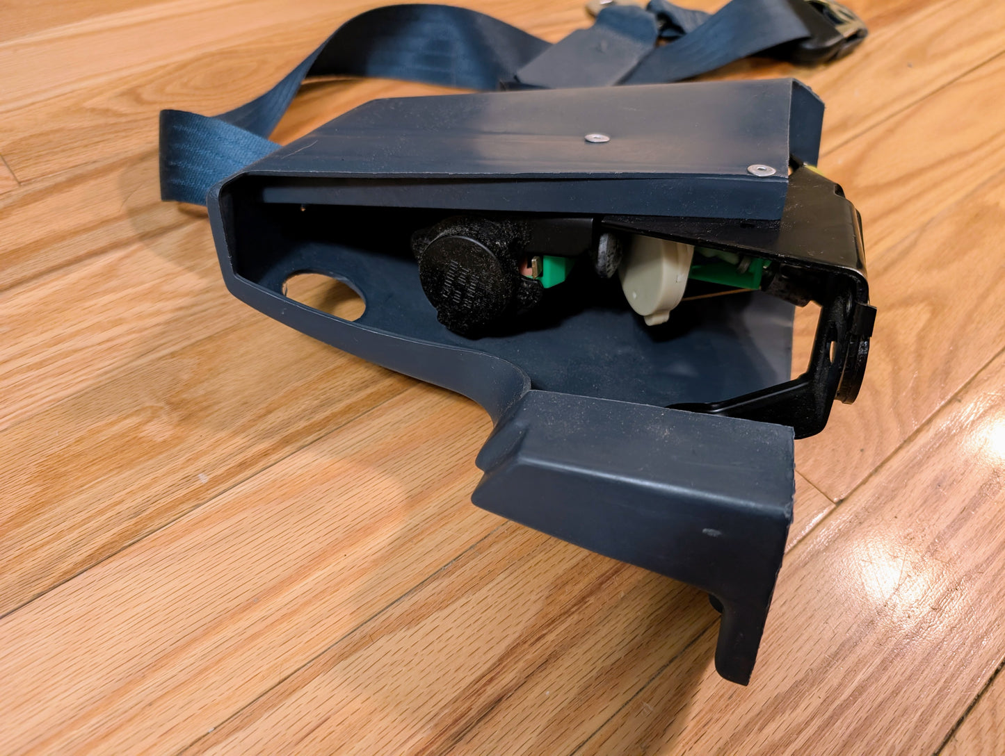 A close-up of a disassembled blue NOS General Motors passenger seat belt retractor for 1982-1993 Chevy S10/GMC S15, showing internal components like a green circuit board and black and white parts on a wooden floor.