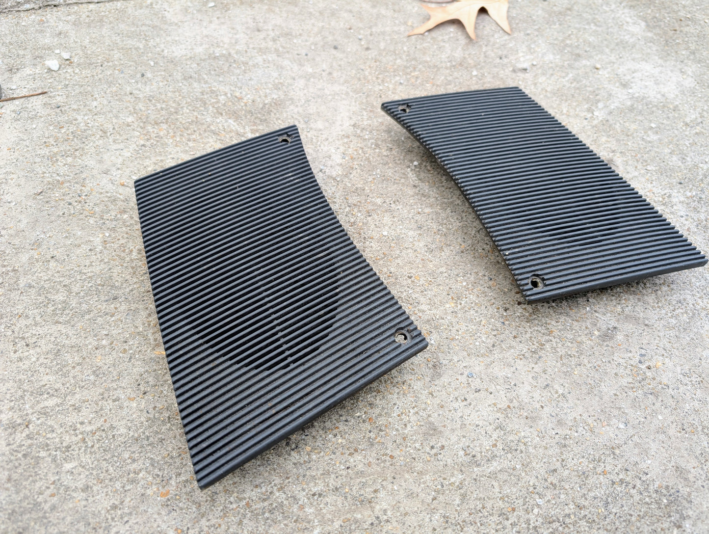 Two charcoal rectangular OEM dash speaker grille covers by General Motors for 1986-1993 Chevrolet S10, Blazer, and more sit on rough concrete with a brown leaf nearby—perfect replacements for your original speaker grills.