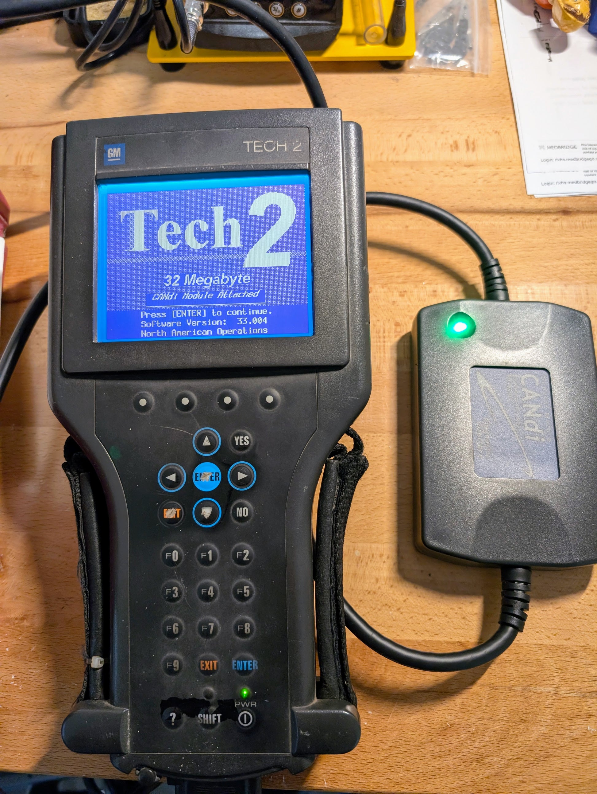 A Vetronix TESTED Tech 2 Automotive Diagnostic Tool & Accessories for 1991-2013 GM Vehicles is shown with a Tech 2, 32 Megabyte screen, connected to a CANdi module with a green light, all placed on a wooden surface.