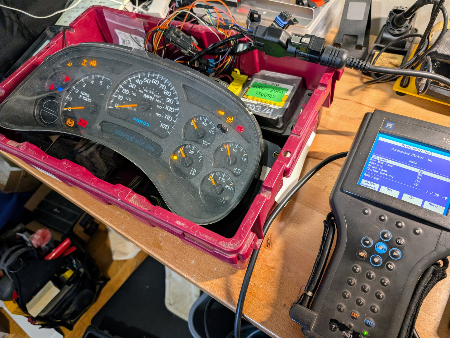 A Vetronix TESTED Tech 2 Automotive Diagnostic Tool & Accessories for 1991-2013 GM Vehicles is connected to a car dashboard instrument cluster in a red bin, surrounded by wires on a cluttered workbench.