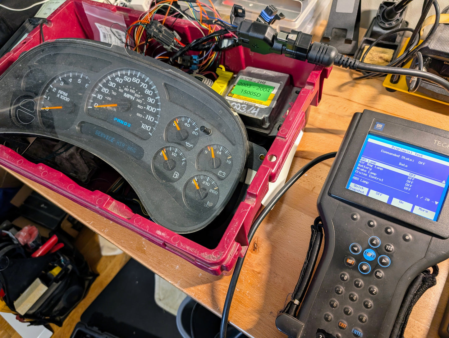 A Vetronix TESTED Tech 2 Automotive Diagnostic Tool & Accessories for 1991-2013 GM vehicles connects by wires to a car instrument cluster on a workbench, showing vehicle data, with various electronic components surrounding them.