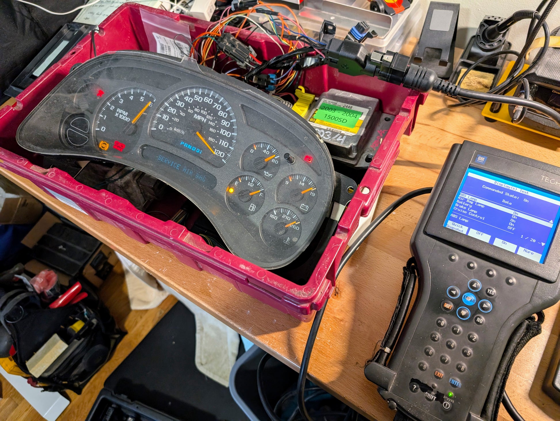 A Vetronix TESTED Tech 2 Automotive Diagnostic Tool & Accessories for 1991-2013 GM Vehicles is connected to a disassembled car instrument cluster on a workbench, amid wires and electronic components in a workshop.