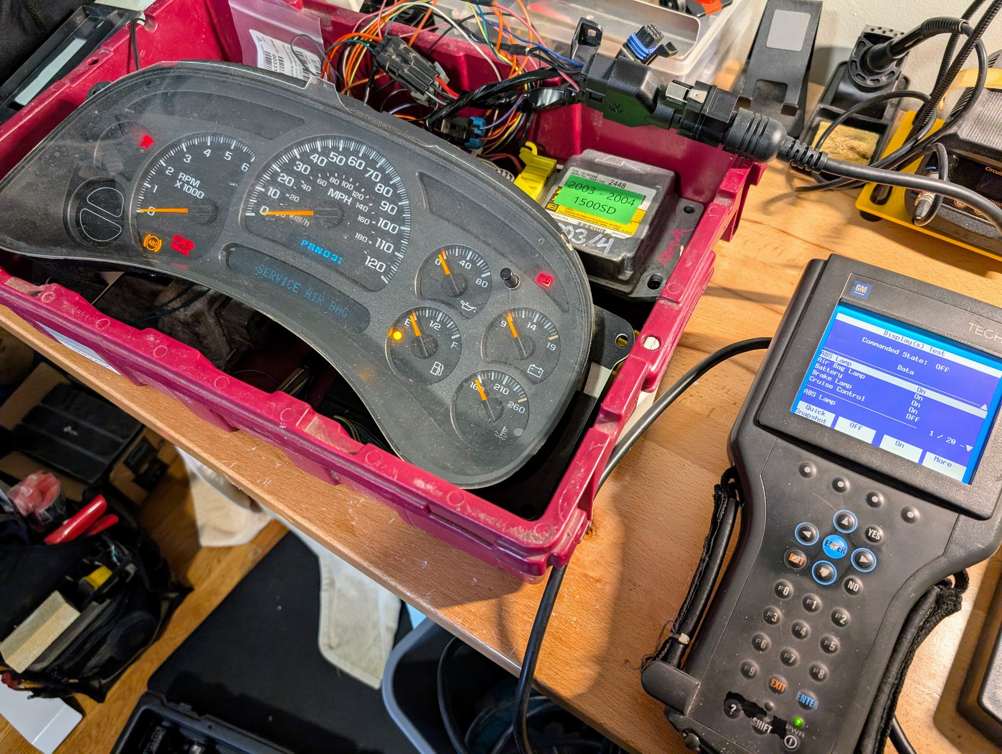 A Vetronix TESTED Tech 2 Automotive Diagnostic Tool & Accessories for 1991-2013 GM Vehicles is connected to a disassembled car dashboard instrument cluster on a cluttered workbench with wires and electronic components.