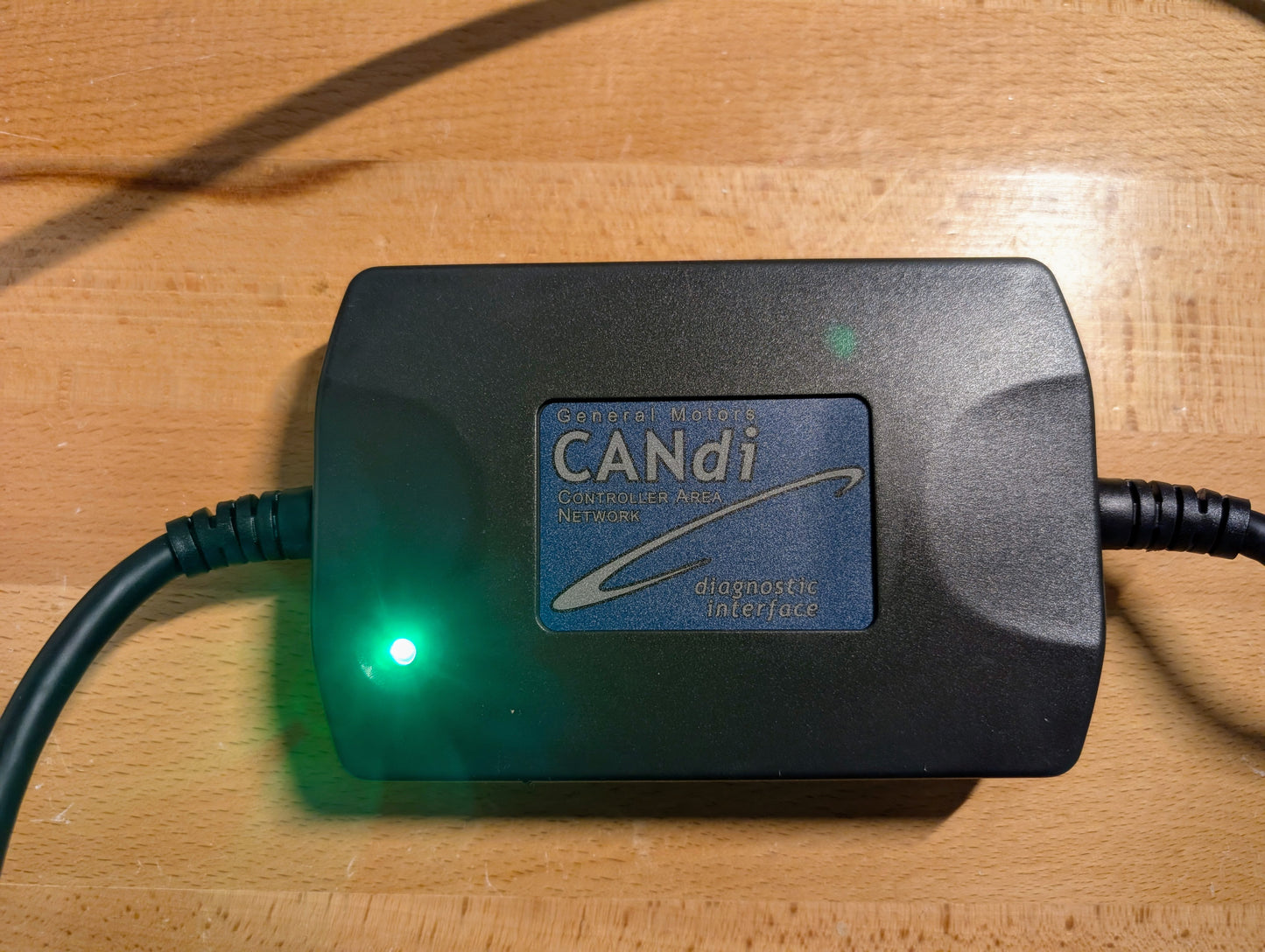 A black Vetronix TESTED Tech 2 Automotive Diagnostic Tool & Accessories for 1991-2013 GM Vehicles, featuring a glowing green indicator light and cables attached on both sides, rests on a wooden surface.