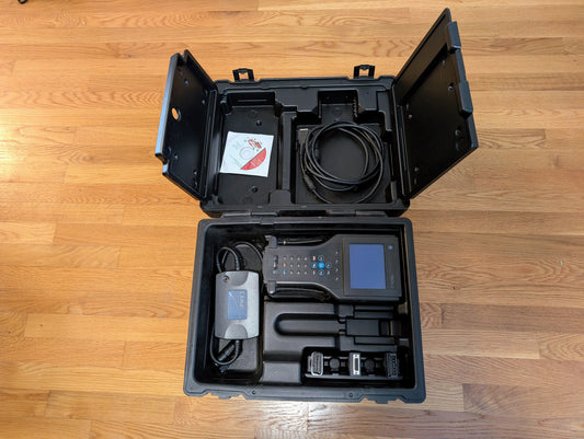 A black plastic case on a wooden floor contains a Vetronix TESTED Tech 2 Automotive Diagnostic Tool & Accessories for 1991-2013 GM vehicles, including cables, power adapter, and CD in sleeve.